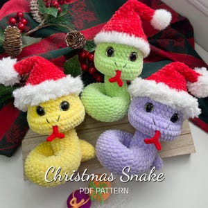 May include: Three crocheted snake toys wearing Santa hats. The snakes are green, yellow, and purple. The hats are red with white trim. The text "Christmas Snake PDF PATTERN" is below the snakes.