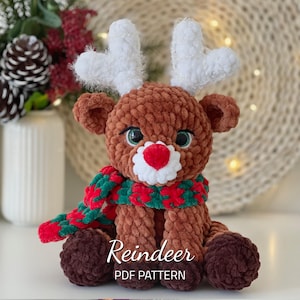 Peut inclure: Un jouet de renne en crochet avec une fourrure brune, des bois blancs, une écharpe verte et rouge et un nez rouge en forme de cœur. Le jouet est assis sur une surface blanche. Patron de renne PDF.