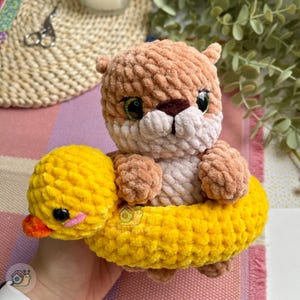 May include: A handmade crochet plush toy featuring a light brown otter with large green eyes and a dark brown nose, sitting inside a bright yellow crochet duck-shaped pool float with a small orange beak and black eyes, crafted from soft yarn.