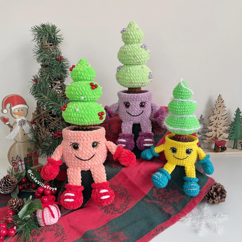 Three crochet Christmas trees in smiling pots are displayed together, each with unique decorations and shades of green, showing a variety of colorful ornaments.