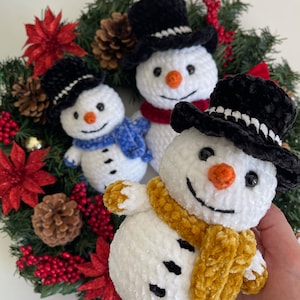 May include: Three crocheted snowmen with black top hats and scarves. The snowmen are arranged on a green wreath with red poinsettias and pine cones.