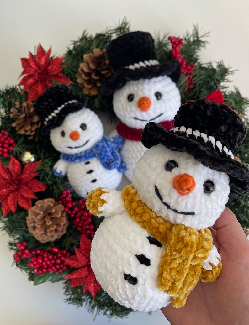 May include: Three crocheted snowmen with black top hats and scarves. The snowmen are arranged on a green wreath with red poinsettias and pine cones.