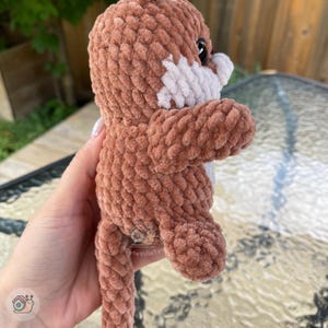 May include: A brown and white crocheted plush toy with a textured, yarn-based surface. The figure features a rounded head, a small snout area with white yarn, a dark eye, and a long tail. The plush is crafted with a consistent, looped stitch pattern.