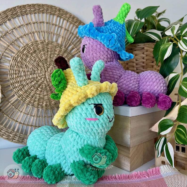 Puede incluir: Dos peluches de oruga hechos a mano con ganchillo. Uno es azul claro con un sombrero amarillo y detalles verdes, el otro es morado con un sombrero azul y detalles verdes. Ambos tienen ojos grandes y est&aacute;n sobre una superficie de madera.