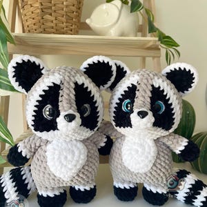 May include: Two handmade crochet raccoon plush toys made from soft, plush yarn. Each raccoon features a grey body, a white chest patch, black eye patches, black ears, and a striped tail. They have large, shiny black eyes and a small black nose.