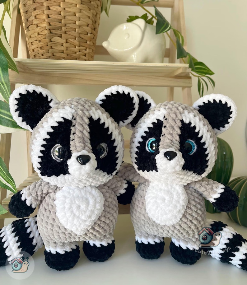 May include: Two handmade crochet raccoon plush toys made from soft, plush yarn. Each raccoon features a grey body, a white chest patch, black eye patches, black ears, and a striped tail. They have large, shiny black eyes and a small black nose.