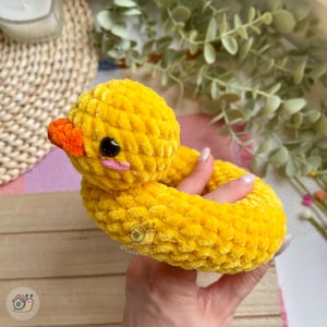 May include: A bright yellow crocheted duck toy with a textured, plush yarn finish. The duck features a small orange beak, black plastic eyes, and pink stitched cheeks. A small logo with a snail graphic and the text CuteVibeToys is visible.