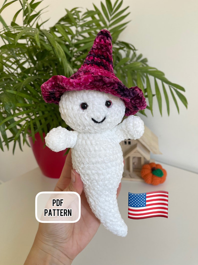 Op de afbeelding: Haakpatroon voor een witte geest met een roze en zwarte heksenhoed. Het patroon is beschikbaar als PDF-download. De afbeelding bevat ook een Amerikaanse vlag.