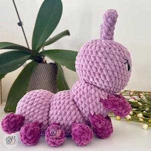 Puede incluir: Un juguete de oruga de peluche hecho a mano en tonos lavanda y morado oscuro. La oruga tiene una cabeza redondeada con peque&ntilde;os ojos negros y una sola antena. Su cuerpo segmentado y sus pies son de un morado m&aacute;s oscuro, creando un juguete suave y caprichoso.