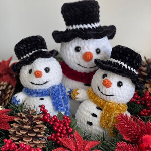 May include: Three crocheted snowmen with black velvet hats and scarves, sitting in a Christmas wreath with red berries, pine cones, and poinsettias.