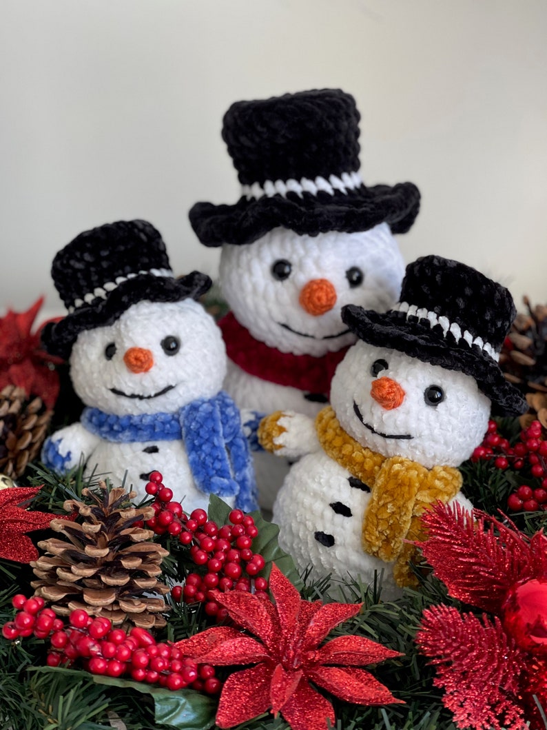 May include: Three crocheted snowmen with black velvet hats and scarves, sitting in a Christmas wreath with red berries, pine cones, and poinsettias.