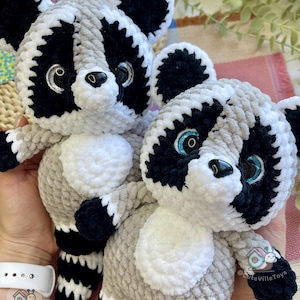 May include: Two plush raccoon toys made of soft, chunky yarn in shades of gray, black, and white. Each raccoon features large, glossy eyes with blue or silver glitter accents, a black nose, and a striped tail. The text @CuteVilleToys is visible.