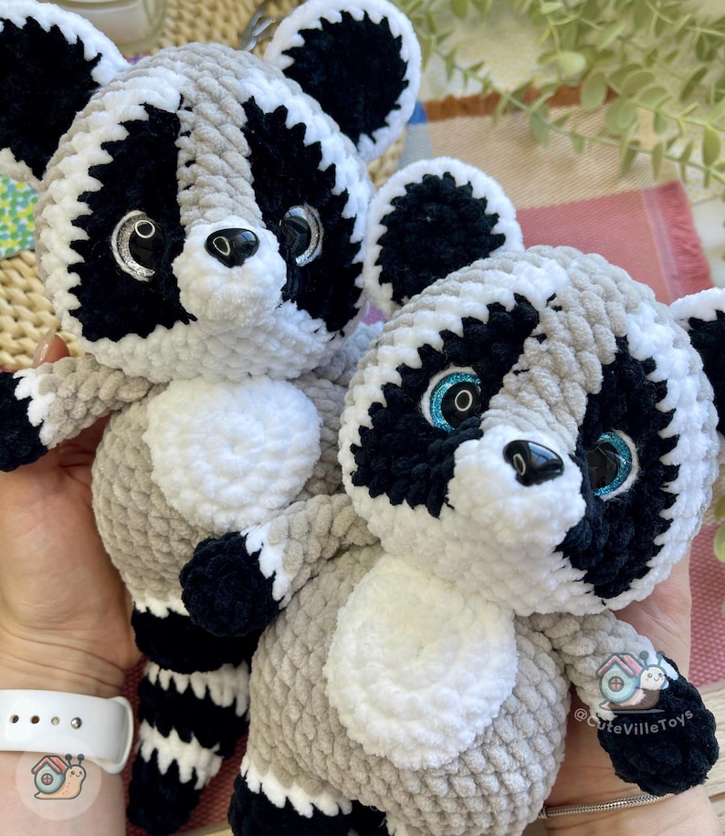 May include: Two plush raccoon toys made of soft, chunky yarn in shades of gray, black, and white. Each raccoon features large, glossy eyes with blue or silver glitter accents, a black nose, and a striped tail. The text @CuteVilleToys is visible.