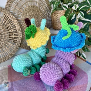 Puede incluir: Dos peluches de oruga hechos a mano con ganchillo. Uno es amarillo y verde azulado, el otro es azul y morado. Cada uno tiene una cabeza en forma de flor con hojas y antenas verdes. Los peluches est&aacute;n sobre una manta estampada.