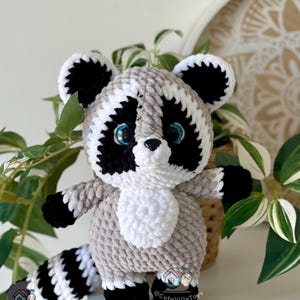 May include: A plush raccoon toy made of soft, textured yarn in shades of gray, black, and white. The raccoon features large, bright blue eyes, a black nose, and a striped tail. A small logo with a snail and the text CuteVilleToys is visible.
