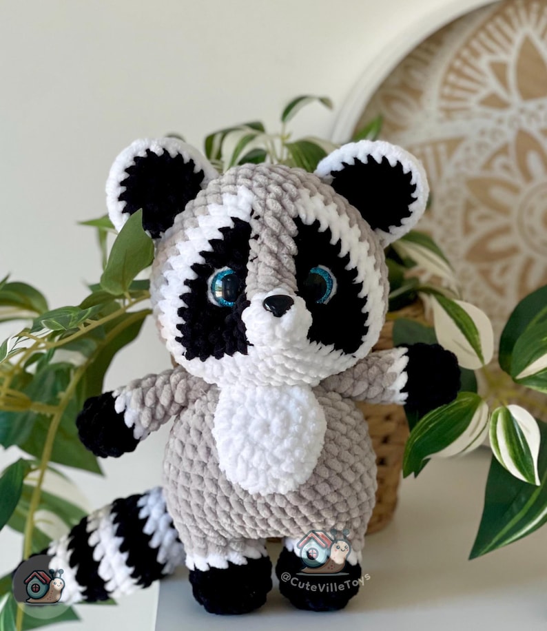 May include: A plush raccoon toy made of soft, textured yarn in shades of gray, black, and white. The raccoon features large, bright blue eyes, a black nose, and a striped tail. A small logo with a snail and the text CuteVilleToys is visible.
