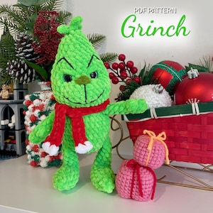 Pode incluir: Um brinquedo de pelúcia Grinch verde feito à mão com um cachecol vermelho, ao lado de duas caixas de presente rosa de crochê. A imagem inclui o texto "PDF Pattern Grinch" em verde. Decorações de Natal no fundo.