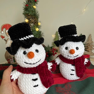 May include: Two crocheted snowmen with black top hats and red scarves. The snowmen are white with black eyes and mouths and orange noses. They are sitting on a red and green plaid surface.