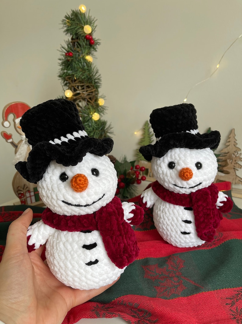 May include: Two crocheted snowmen with black top hats and red scarves. The snowmen are white with black eyes and mouths and orange noses. They are sitting on a red and green plaid surface.