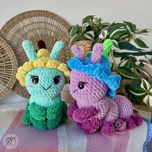 Puede incluir: Dos peluches de oruga hechos a mano con ganchillo. Uno es turquesa, verde y amarillo, el otro es morado, azul y verde. Ambos tienen caras sonrientes, ojos grandes y est&aacute;n sentados sobre una manta estampada. Los juguetes son caprichosos y coloridos.