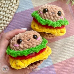 Pode incluir: Dois brinquedos de hambúrguer de pelúcia feitos de fio macio e texturizado. Cada um apresenta um pão bege, uma camada de alface verde brilhante, uma fatia de tomate vermelho e um pão inferior amarelo. Têm olhos de plástico preto e bocas rosas.