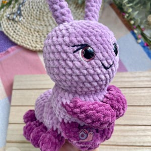 Puede incluir: Un juguete de oruga de peluche hecho a mano en tonos de morado y magenta. La oruga tiene ojos grandes y expresivos con acentos rosados y una boca sonriente. El juguete est&aacute; hecho de un material suave y texturizado, y el dise&ntilde;o general es caprichoso y lindo.