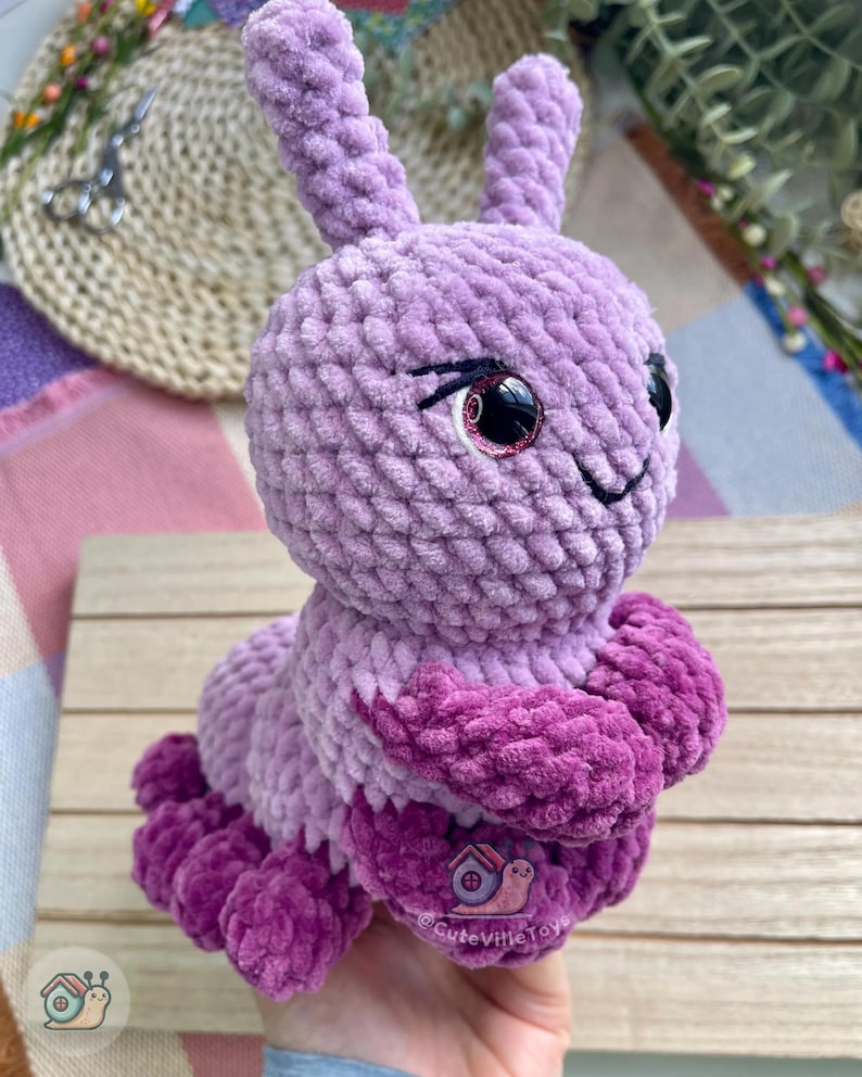 Puede incluir: Un juguete de oruga de peluche hecho a mano en tonos de morado y magenta. La oruga tiene ojos grandes y expresivos con acentos rosados y una boca sonriente. El juguete est&aacute; hecho de un material suave y texturizado, y el dise&ntilde;o general es caprichoso y lindo.