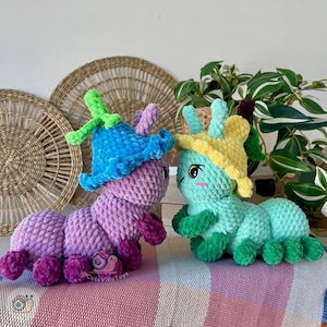 Puede incluir: Dos juguetes de peluche con forma de oruga. Uno es morado con un sombrero azul, el otro es verde azulado con un sombrero amarillo. Ambos tienen caras bonitas y est&aacute;n sobre una manta a rayas. El fondo incluye decoraci&oacute;n de pared tejida y plantas.