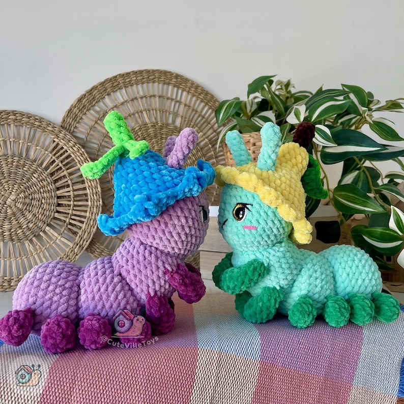 Puede incluir: Dos juguetes de peluche con forma de oruga. Uno es morado con un sombrero azul, el otro es verde azulado con un sombrero amarillo. Ambos tienen caras bonitas y est&aacute;n sobre una manta a rayas. El fondo incluye decoraci&oacute;n de pared tejida y plantas.