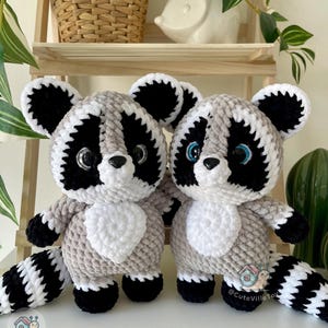 May include: Two plush raccoon toys crafted from soft, textured yarn in shades of gray, black, and white. Each raccoon features large, glossy black eyes, a small black nose, and a striped tail. The toys have white chest patches and ears.