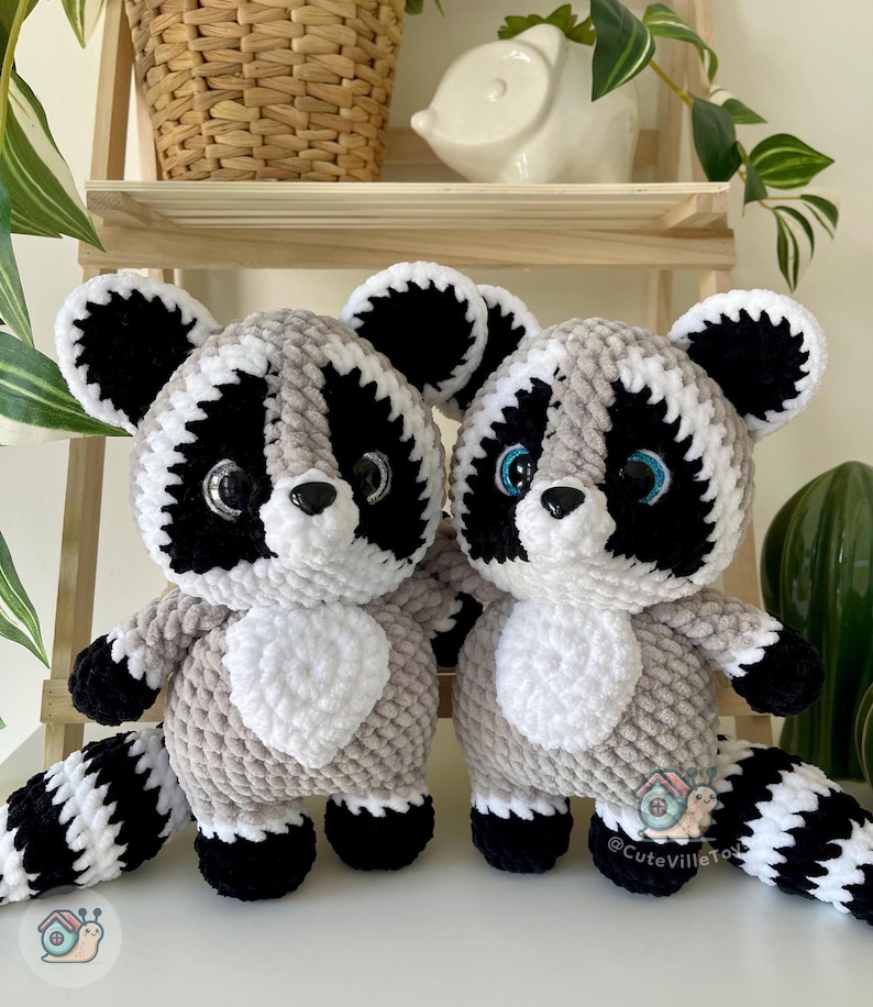 May include: Two plush raccoon toys crafted from soft, textured yarn in shades of gray, black, and white. Each raccoon features large, glossy black eyes, a small black nose, and a striped tail. The toys have white chest patches and ears.