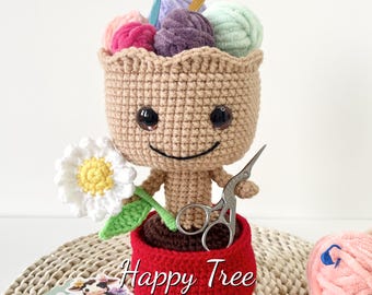 May include: A crocheted amigurumi toy shaped like a tree with a smiling face. The tree is holding a pair of scissors and has a pot with a red rim. The tree is surrounded by yarn balls and a daisy.