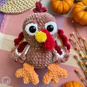 May include: A plush turkey toy made of brown, soft, textured yarn. It features large black plastic eyes with white yarn circles, a bright yellow beak, and a red wattle. The turkey has orange feet and a tail with layers of red, white, and brown yarn.