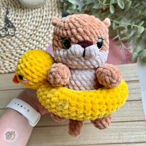 May include: A crocheted light brown otter plush toy with dark green eyes and a white muzzle, positioned inside a bright yellow crocheted duck-shaped floatie. The duck floatie features a small orange beak and black eyes, crafted from soft yarn.