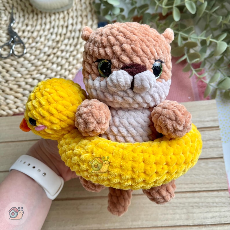 May include: A crocheted light brown otter plush toy with dark green eyes and a white muzzle, positioned inside a bright yellow crocheted duck-shaped floatie. The duck floatie features a small orange beak and black eyes, crafted from soft yarn.