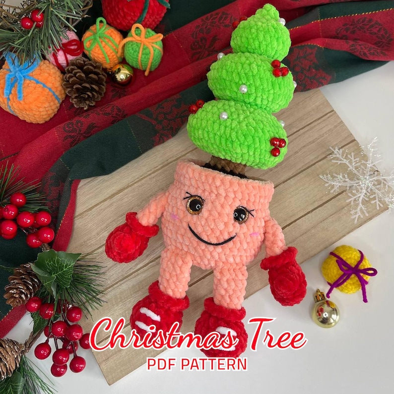 K&ouml;nnte beinhalten: Ein geh&auml;keltes Weihnachtsbaumspielzeug in einem rosa Topf mit einem l&auml;chelnden Gesicht und roten F&auml;ustlingen. Der Baum ist gr&uuml;n mit roten Beeren und wei&szlig;en Perlen. Das Spielzeug befindet sich auf einer Holzoberfl&auml;che mit einem rot-gr&uuml;nen karierten Stoff im Hintergrund. Weihnachtsbaum-PDF-Muster.