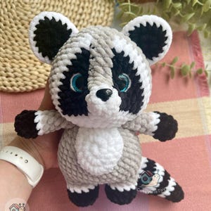 May include: A plush raccoon toy made of soft, textured yarn in shades of gray, black, and white. The raccoon features large, bright blue eyes, a black nose, and a striped tail. A small logo with a snail and the text CuteVilleToys is visible.