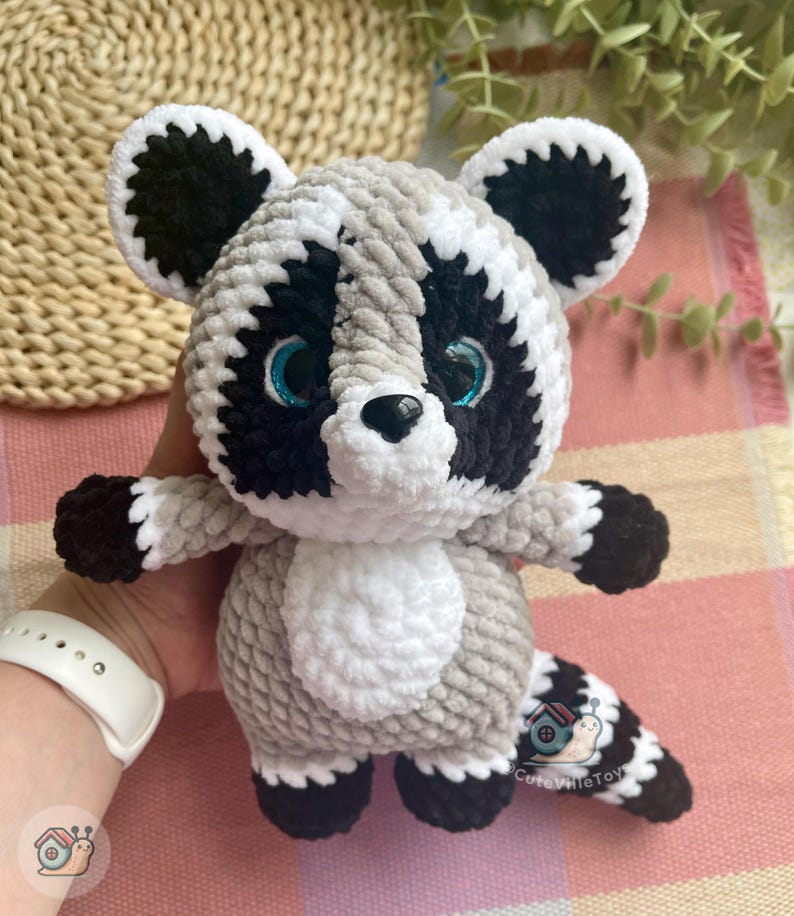 May include: A plush raccoon toy made of soft, textured yarn in shades of gray, black, and white. The raccoon features large, bright blue eyes, a black nose, and a striped tail. A small logo with a snail and the text CuteVilleToys is visible.