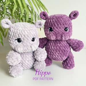 Op de afbeelding: Twee gehaakte nijlpaard knuffels, één in lichtpaars en één in donkerpaars. De knuffels hebben zwarte ogen en zitten op een wit oppervlak. De tekst "Hippo PDF Pattern" staat onder de knuffels.