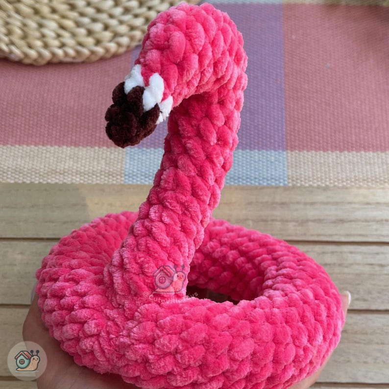May include: A bright pink crocheted flamingo figure with a long, curved neck. The beak is dark brown with a small white accent near the base. The texture is soft and plush, created with thick yarn stitches. A small logo is visible on the neck.
