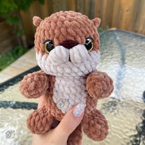 May include: A small, plush, crocheted otter toy made from soft, brown and cream-colored yarn. The otter features large, glossy black eyes with gold-colored rims, a dark brown nose, and a light cream-colored belly patch with a small snail logo.