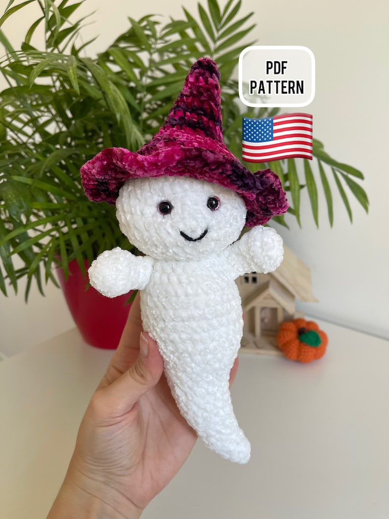 Op de afbeelding: Een witte gehaakte geest die een roze en zwarte fluwelen heksenhoed draagt. De geest heeft zwarte ogen en een glimlachende mond. De afbeelding bevat de tekst "PDF PATTERN" en een kleine Amerikaanse vlag.