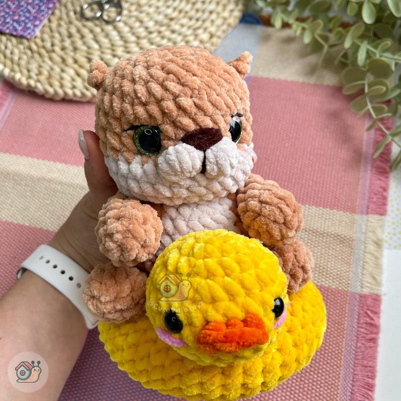 May include: A plush crochet toy featuring a light brown, fuzzy animal with dark eyes and a white muzzle, sitting atop a bright yellow, round crochet duckling with a small orange beak and pink cheeks, crafted from soft, textured yarn material.