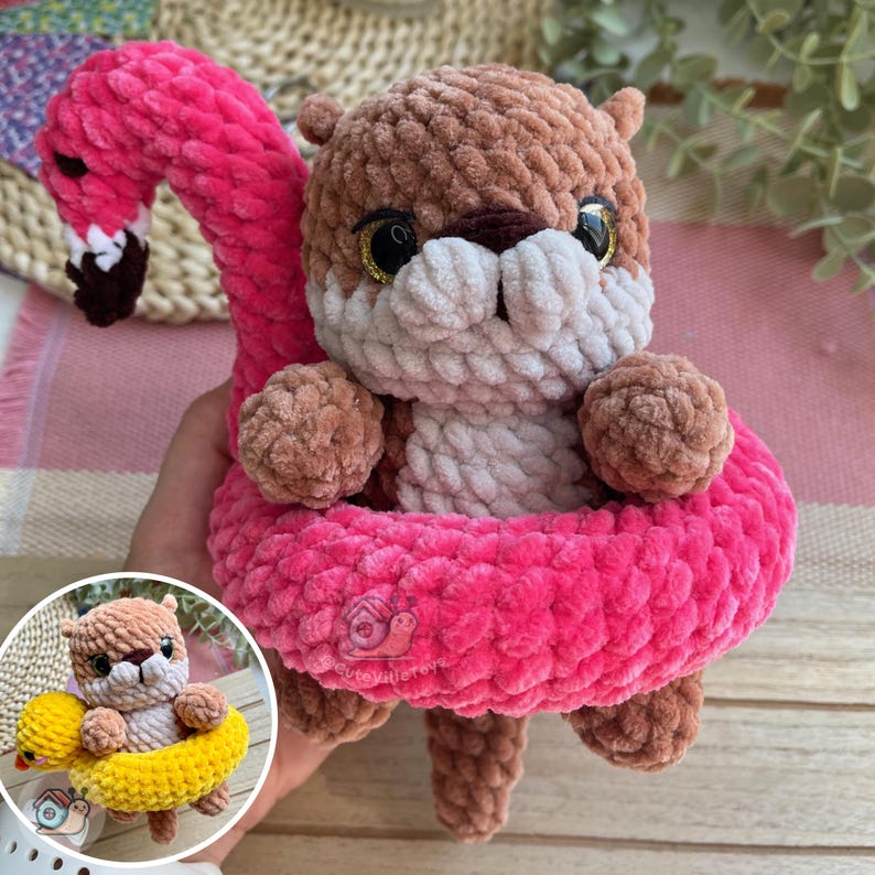 May include: A brown and white crocheted otter plush toy sitting inside a bright pink flamingo-shaped pool float. The otter has large black eyes and a soft, textured yarn finish. A small logo is visible on the front of the pink float.
