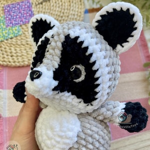 May include: A plush raccoon toy crafted from soft, textured yarn in shades of gray, black, and white. The figure features a distinct black mask around its eyes, white facial accents, rounded ears with black centers, and a small black nose.