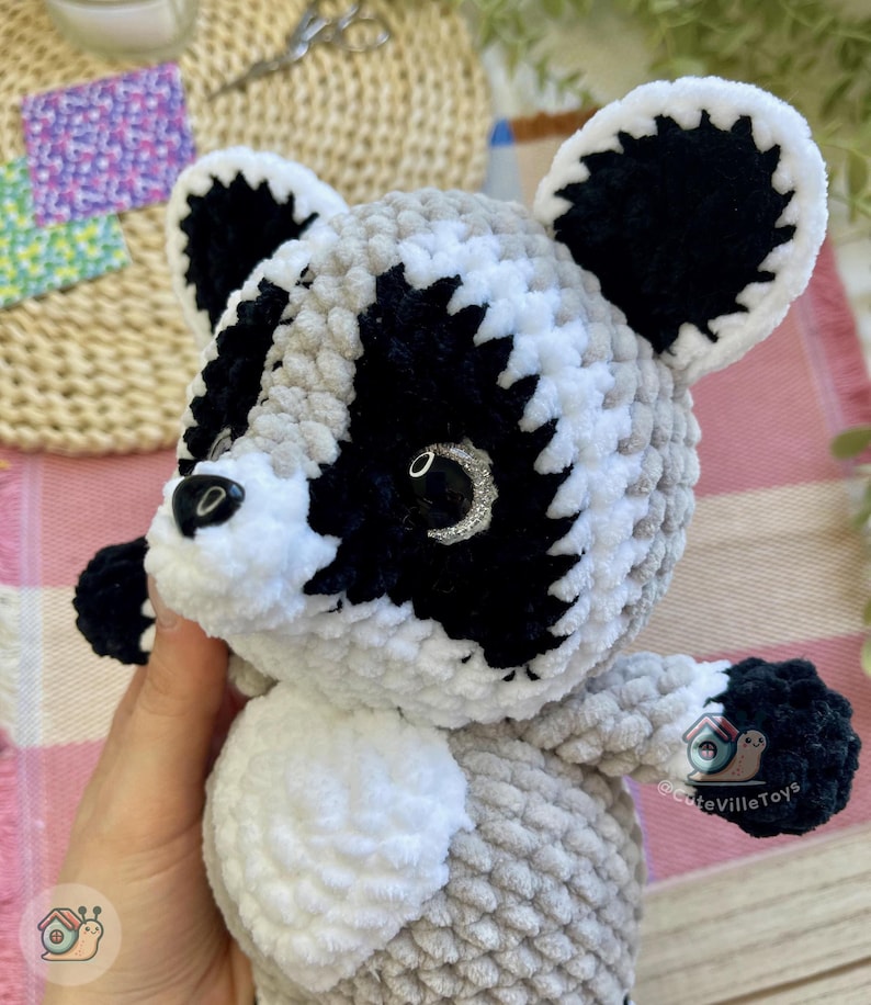 May include: A plush raccoon toy crafted from soft, textured yarn in shades of gray, black, and white. The figure features a distinct black mask around its eyes, white facial accents, rounded ears with black centers, and a small black nose.