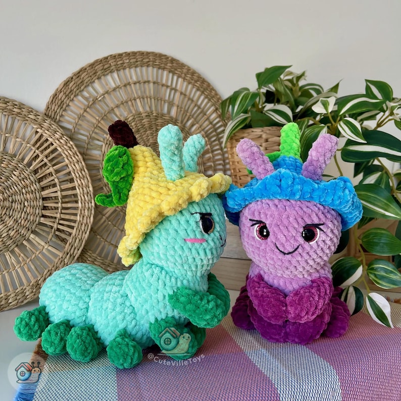 Puede incluir: Dos juguetes de oruga hechos a mano con ganchillo. Uno es azul claro con un sombrero amarillo y detalles verdes. El otro es morado con un sombrero azul y detalles rosas. Ambos tienen caras sonrientes y est&aacute;n sobre una superficie a rayas moradas y blancas.