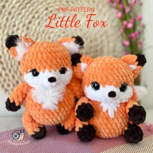 May include: Two crocheted fox plushies, one larger and one smaller, both with orange fur, white accents on their chests, and black noses and feet. The plushies are sitting on a pink and white patterned surface. The text "PDF PATTERN Little Fox" is visible above the plushies.