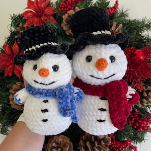 May include: Two white crocheted snowmen with black top hats and scarves. The snowman on the left has a blue scarf and the snowman on the right has a red scarf. The snowmen are sitting in a Christmas wreath with red berries and pine needles.