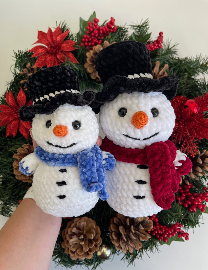 May include: Two white crocheted snowmen with black top hats and scarves. The snowman on the left has a blue scarf and the snowman on the right has a red scarf. The snowmen are sitting in a Christmas wreath with red berries and pine needles.